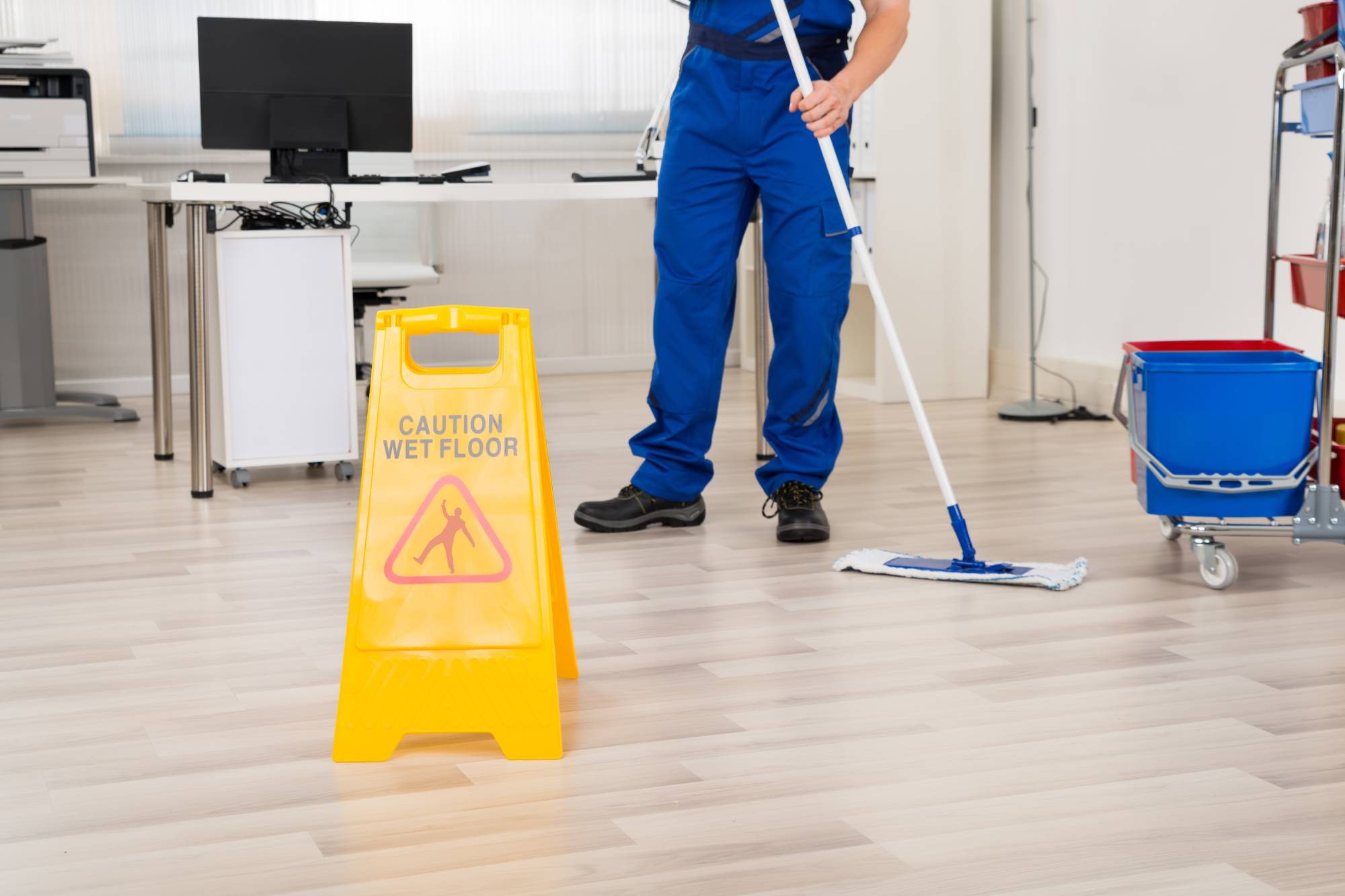 Eine Reinigungsfachkraft in blauer Arbeitskleidung steht in einem Büroraum und reinigt Laminatboden mit einem Wischmopp. Ein gelbes Warnschild im Vordergrund warnt vor Rutschgefahr.