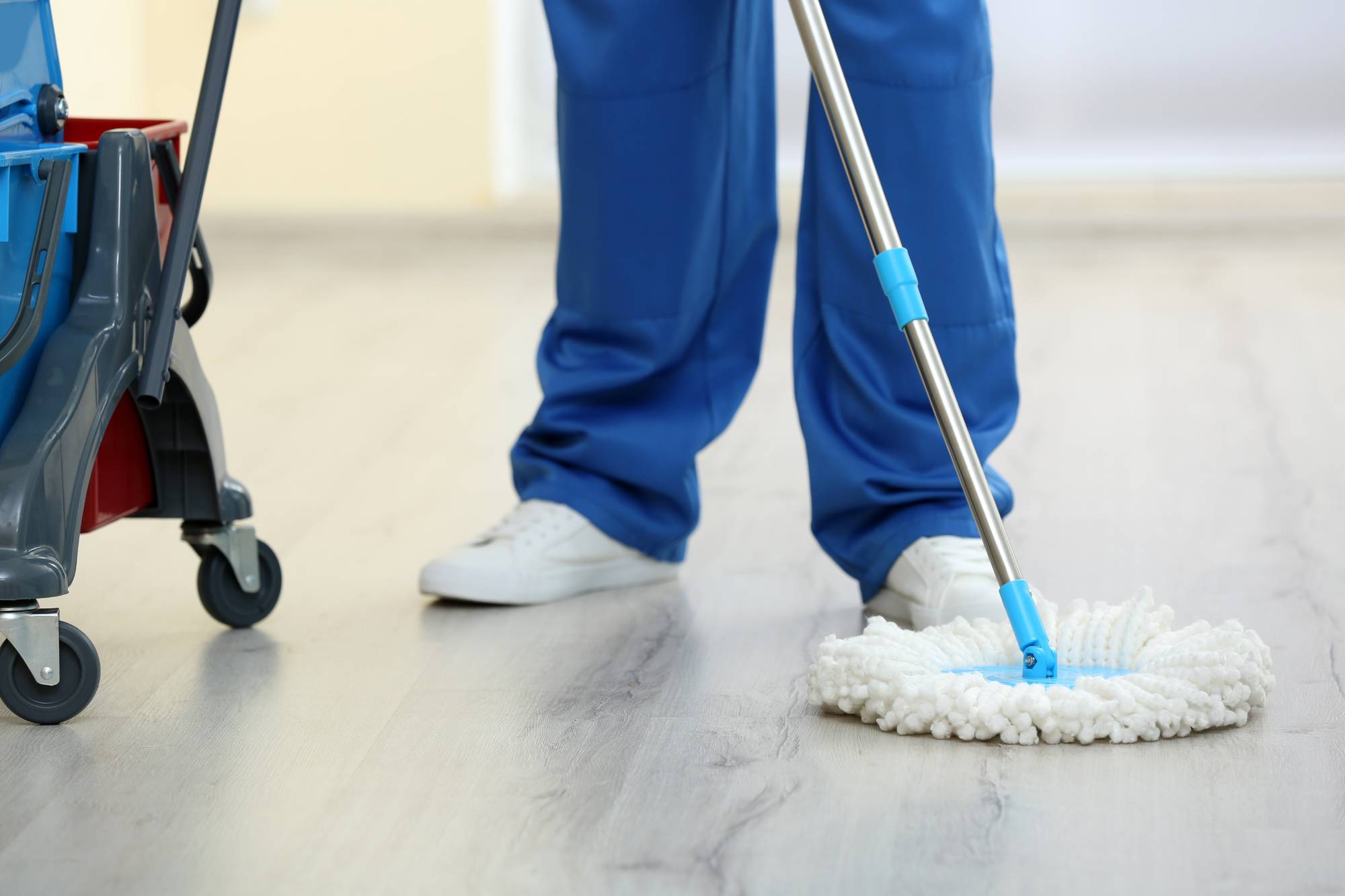 Eine Reinigungsfachkraft mit blauer Hose und weißen Schuhen fährt mit einem runden Mikrofaser-Mop über einen glatten Holzboden